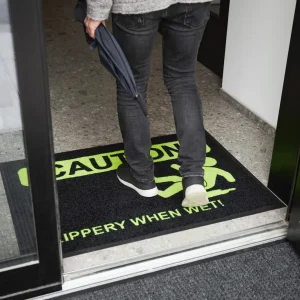 Človek stojaci na čiernej rohoži JET PRINT HI-VIS so žltým upozornením CAUTION a symbolom pošmyknutia pri dverách