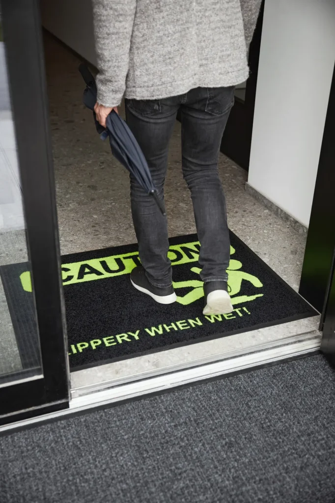 Človek stojaci na čiernej rohoži JET PRINT HI-VIS so žltým upozornením CAUTION a symbolom pošmyknutia pri dverách