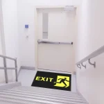 Čierna rohož JET PRINT HI-VIS so žltým nápisom EXIT a symbolom východu pred dverami na schodiskovej podeste