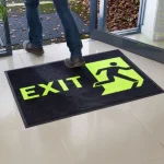 Čierna rohož JET PRINT HI-VIS so žltým nápisom EXIT a symbolom východu pri otvorených presklených dverách