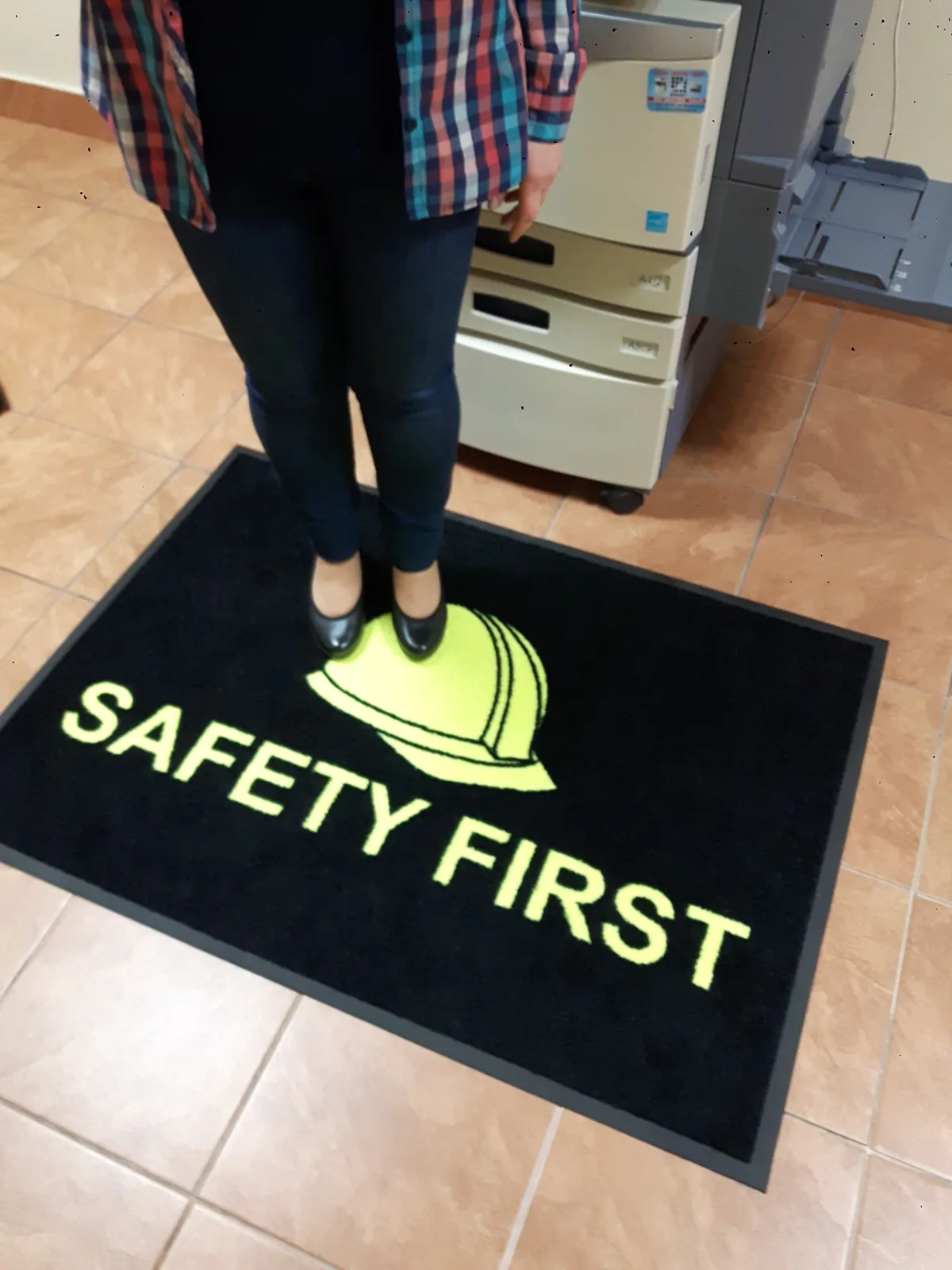 Človek stojaci na čiernej rohoži JET PRINT HI-VIS so žltou prilbou a nápisom SAFETY FIRST pri zariadení