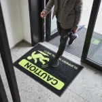 Rohož JET PRINT HI-VIS s nápismi CAUTION a SLIPPERY WHEN WET pod človekom pri presklenom vstupe
