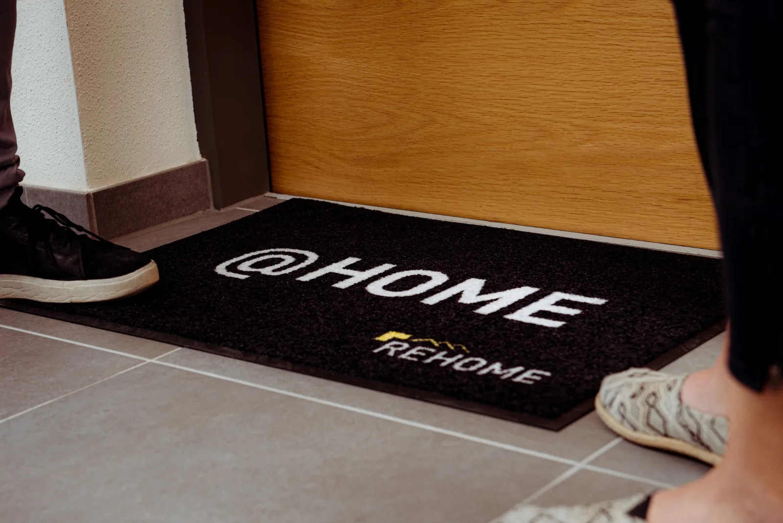 Čierna rohož JET PRINT PATIO s bielym nápisom @HOME a logom REHOME pred dverami