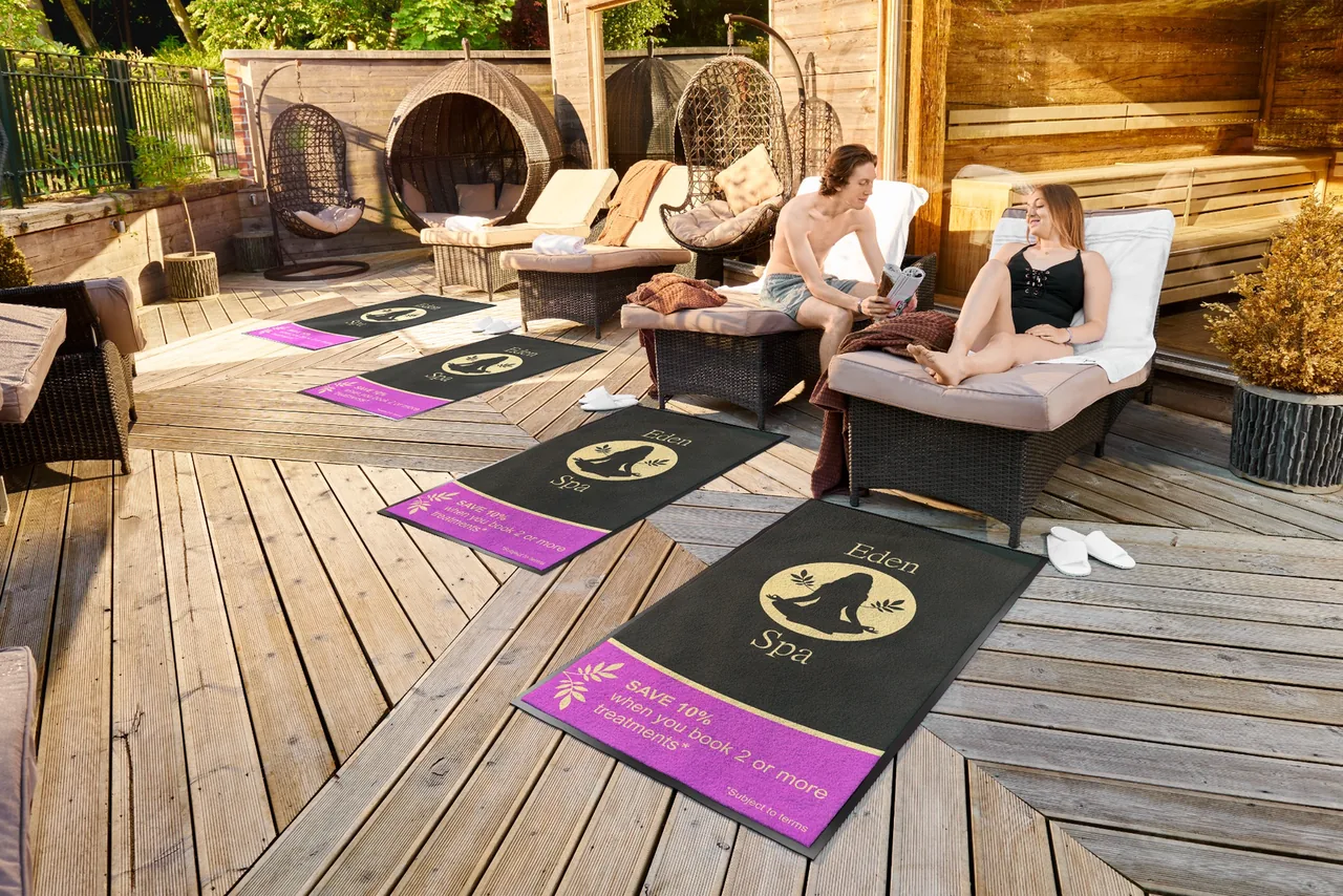 Čierne a fialové rohože JET PRINT PATIO s logom pred oddychovými ležadlami na drevenej terase